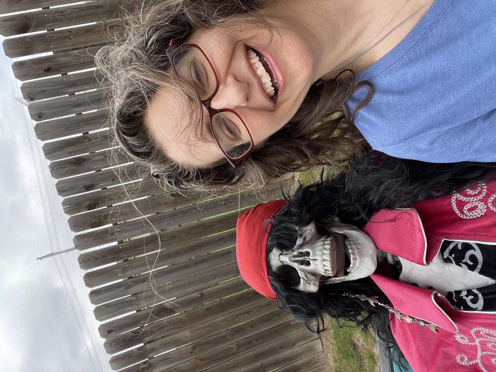 Selfie with The Dead Pirate Robert. Image of me, a woman with red glasses and curly hair laughing next to her plastic skeleton dressed as a pirate.