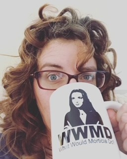 Woman with curly hair wearing glasses and drinking from a mug that has a picture of Morticia Addams on it and reads "WWMD What would Morticia Do?"