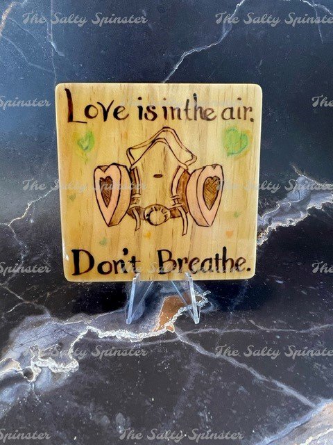 Photo of four inch wooden tile square on a clear acrylic stand infront of a black marble background. Wood has been burned with the words "Love is in the air. Don't Breathe" and has an image of a respirator breathing mask with heart shaped filters. Watercolor accents have been added.