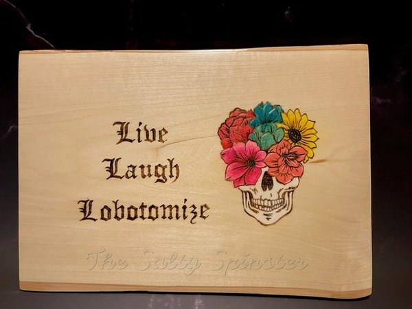 Wood wall plaque featuring gothic lettering that reads "live laugh lobotmize" and includes skull with floral arrangement. Lettering and outlines are burned into the wood. Flowers and skull have watercolor accents.