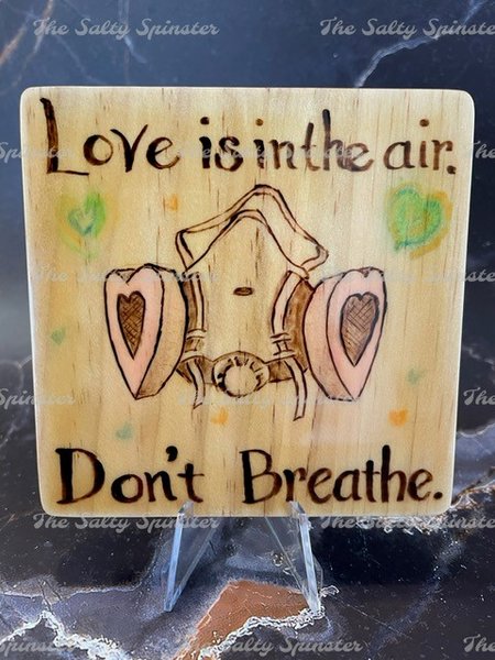 Close up photo of four inch wooden tile square. Wood has been burned with the words "Love is in the air. Don't Breathe" and has an image of a respirator breathing mask with heart shaped filters. Watercolor accents have been added.